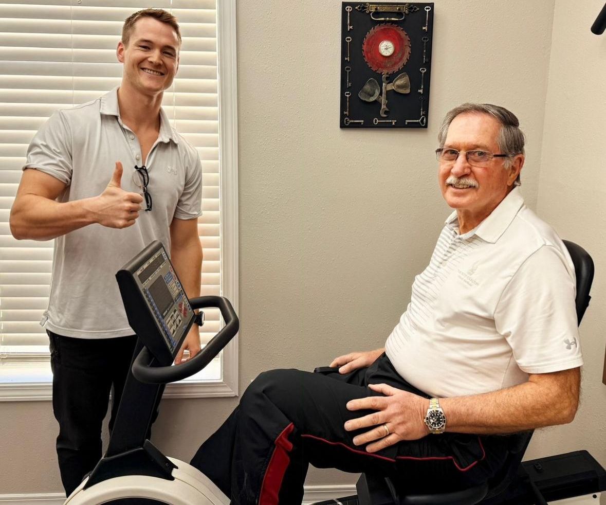 Dr. Austin Baskin giving a thumbs-up beside a smiling older patient seated on a recumbent exercise bike